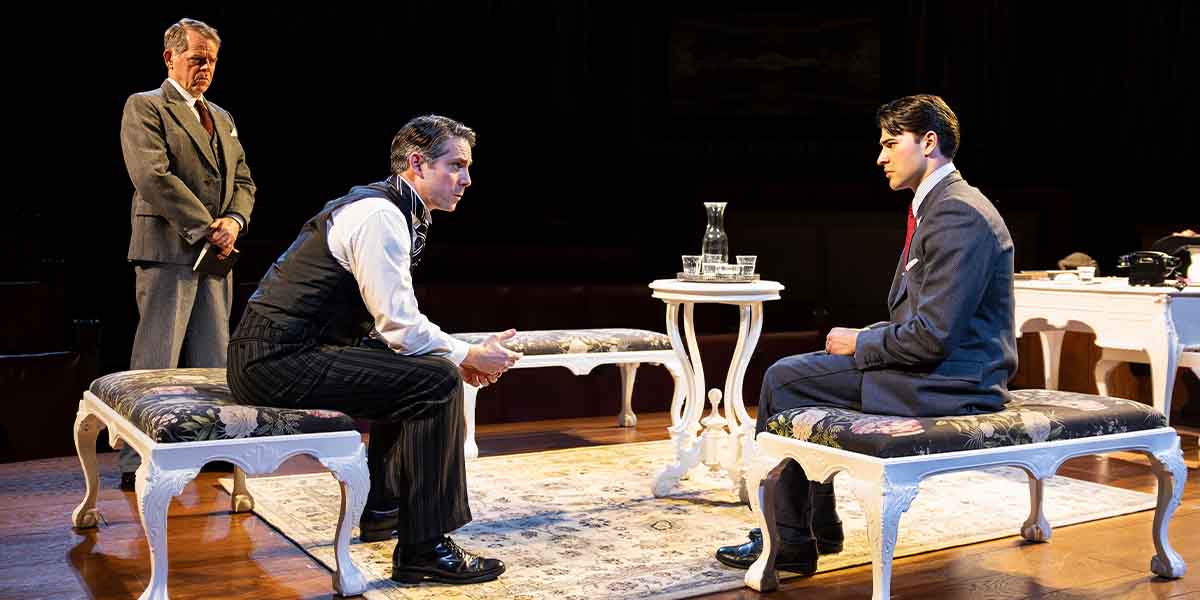 Three people are seated in a courtroom setting, engaged in a serious discussion, with a table between them.