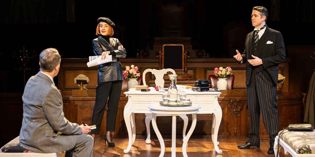 A person in a black outfit and beret stands confidently, while another gestures, seated nearby.