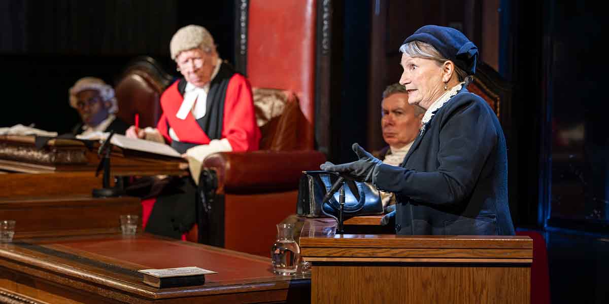 A person in a dark outfit gestures while speaking at a courtroom, with judges and lawyers present.