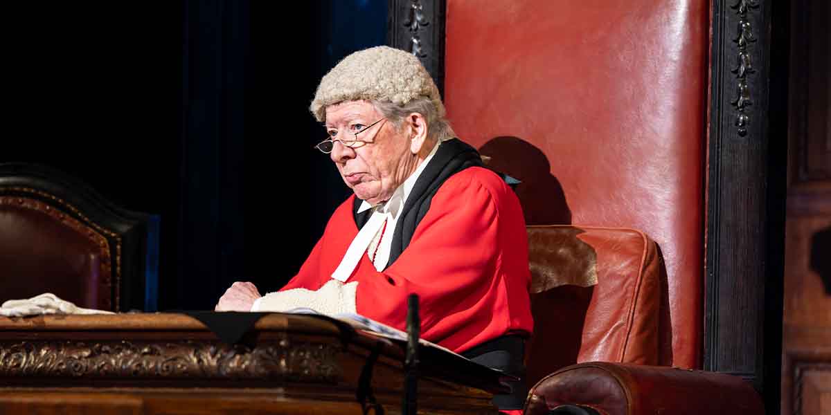 A judge in traditional robes and wig sits at a wooden desk, looking thoughtfully ahead in a courtroom.
