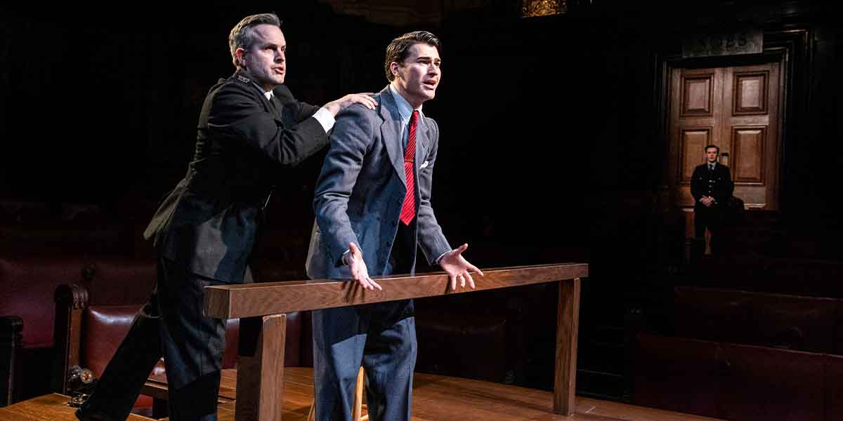 A person in a suit gestures dramatically at a wooden bench while another stands behind.