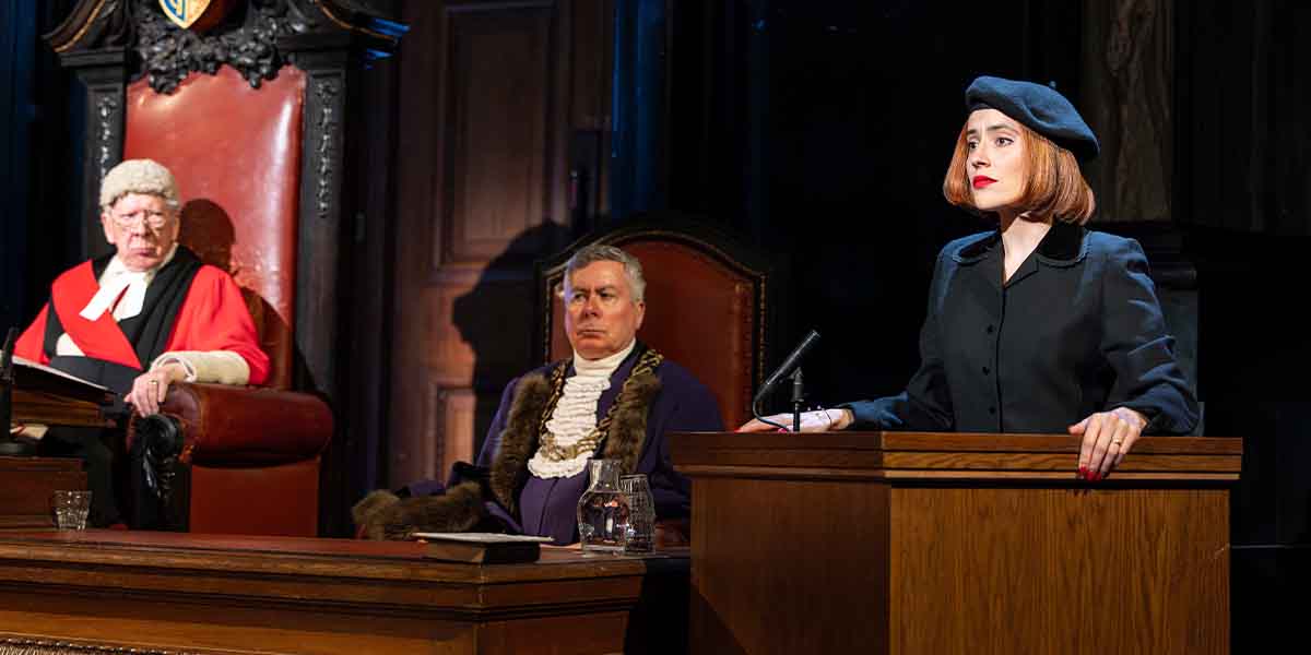 A person in a black outfit and beret stands at a wooden lectern, addressing two others seated in court.
