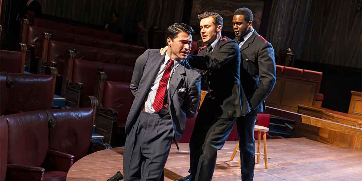 Three people in formal attire perform a dramatic scene in a courtroom setting, with wooden benches visible.
