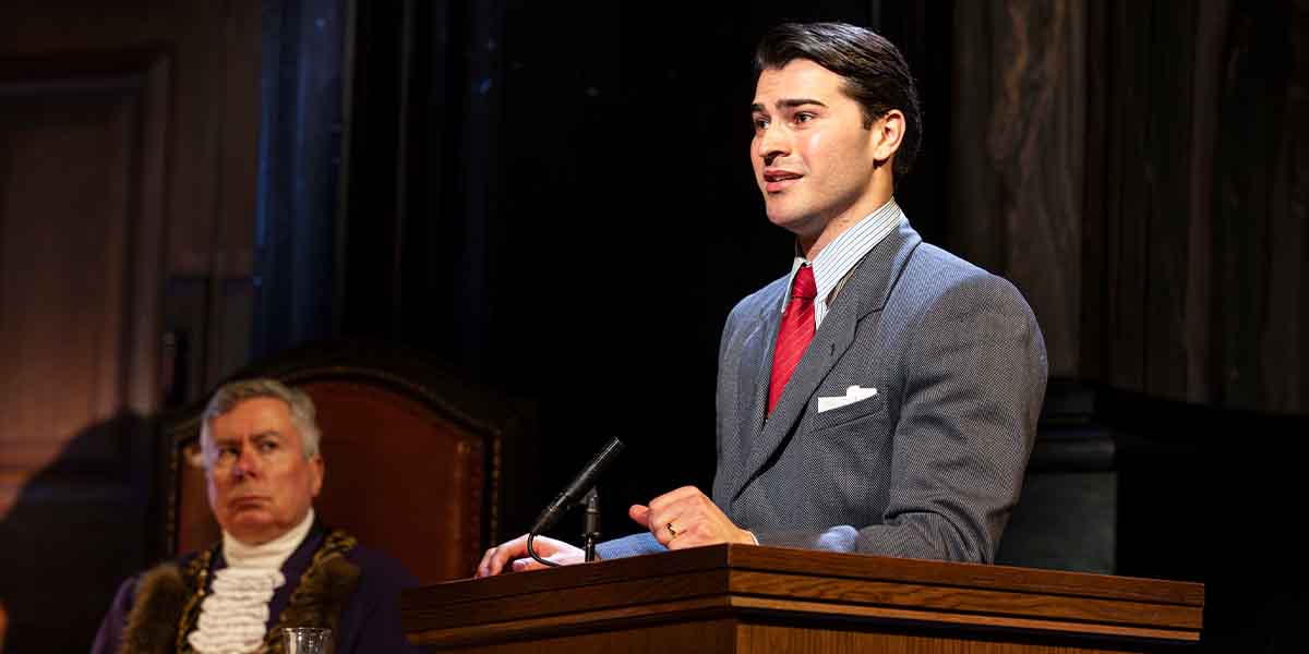 A person in a grey suit and red tie speaks at a wooden lectern, while a judge listens.