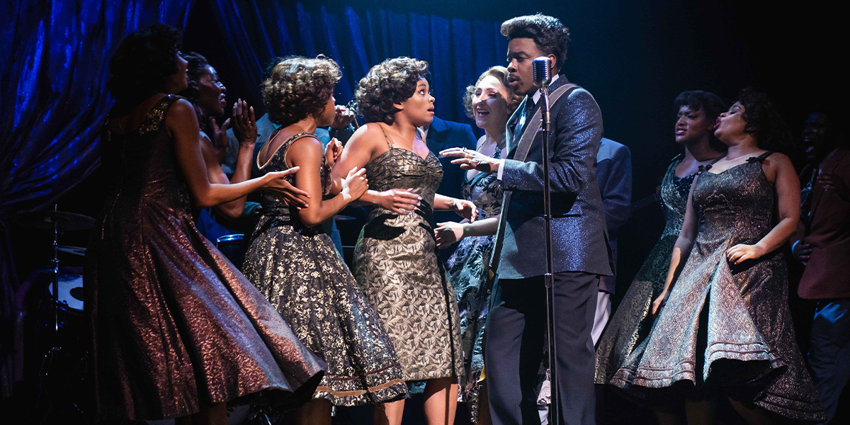 Performers on stage, with one singing into a vintage microphone and others providing vocal support, all dressed in elegant vintage-style costumes.