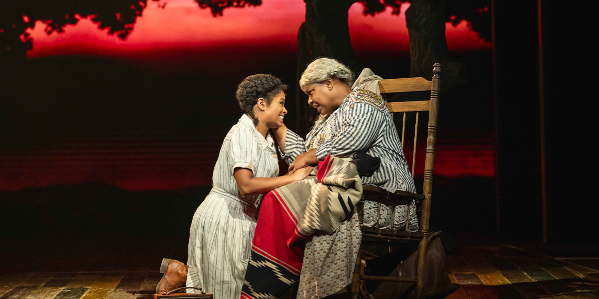 Two actors on stage, one sitting with a blanket on their lap and the other leaning over with hands on their knee, sharing a joyful moment against a backdrop simulating a sunset.