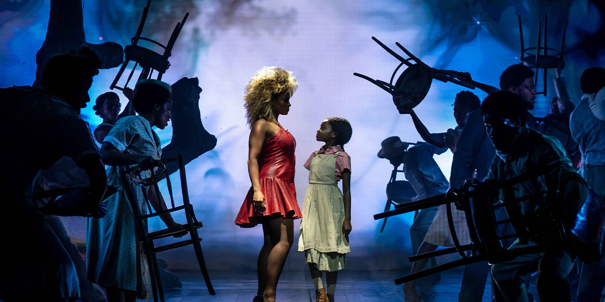 Two actors on stage in a theatrical production, one dressed in a red dress and the other in a patterned shirt and overalls, interacting under dramatic lighting with shadowy figures in the background.