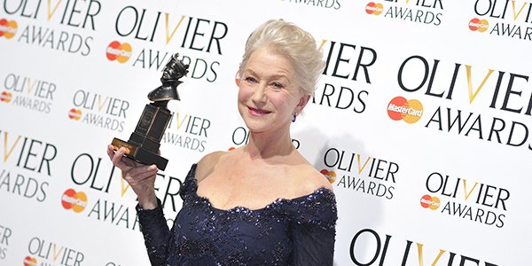 Helen Mirren smiles while holding an award, wearing a navy blue gown against an Olivier Awards backdrop.