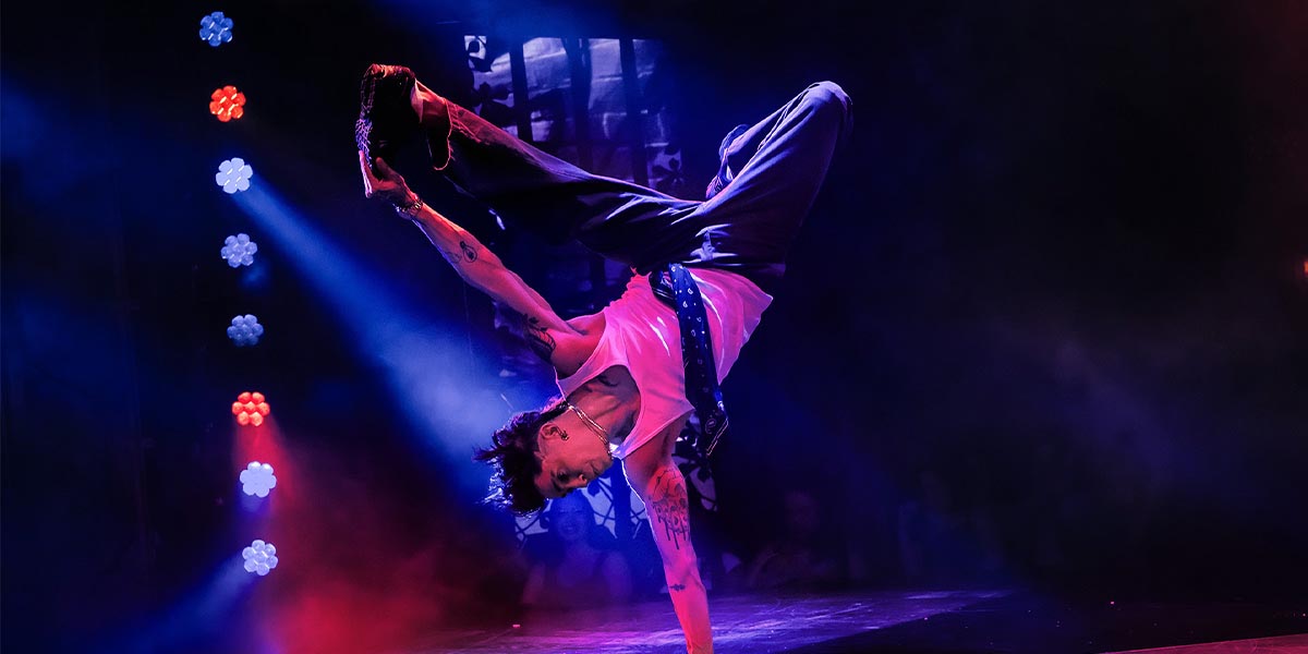 Danny Kaan performs a handstand on stage, illuminated by colourful lights against a smoky backdrop.