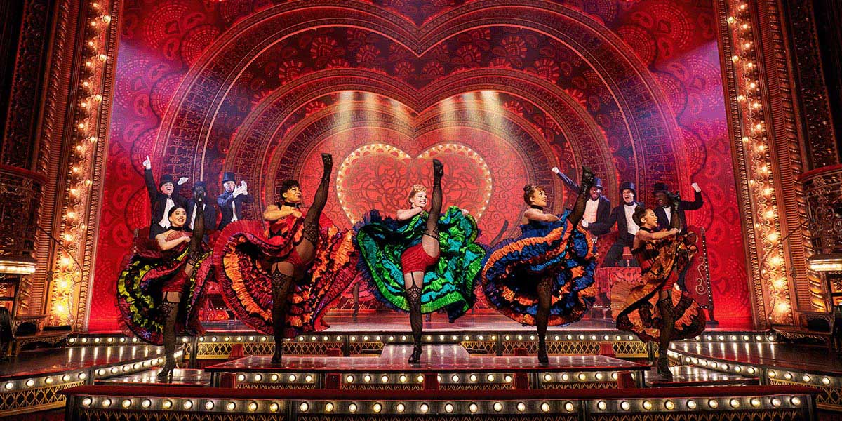 Matt Crockett performs on stage in a vibrant costume, surrounded by colourful set designs from Moulin Rouge! The Musical.
