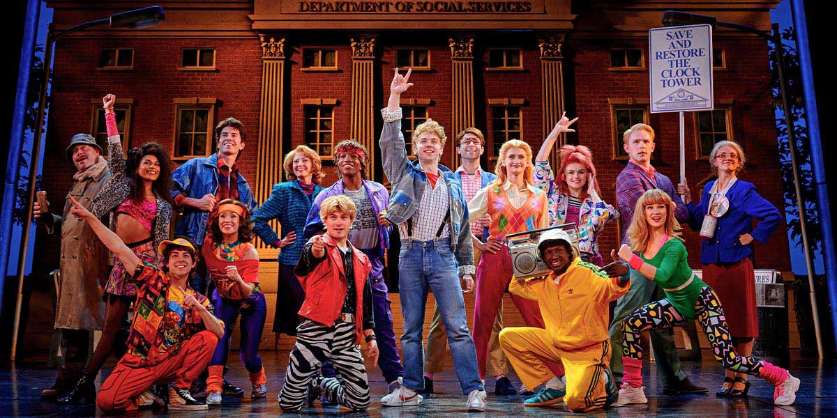 Caden Brauch as Marty McFly, surrounded by cast members, performing energetically on stage with vibrant costumes.