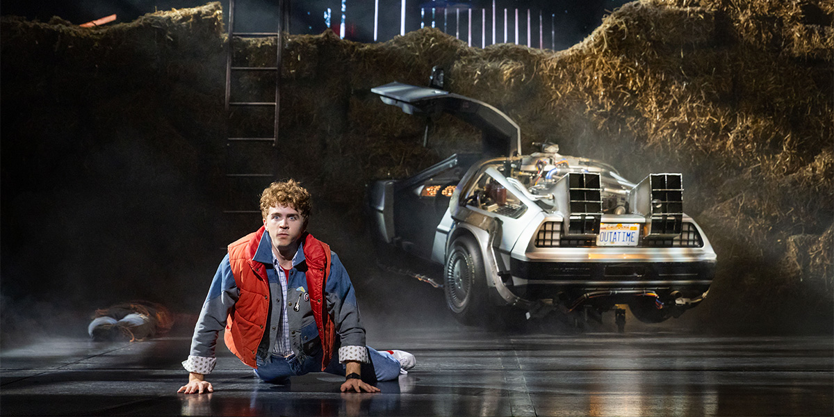 Caden Brauch as Marty McFly, wearing a red vest and jeans, poses energetically on stage with dramatic lighting.