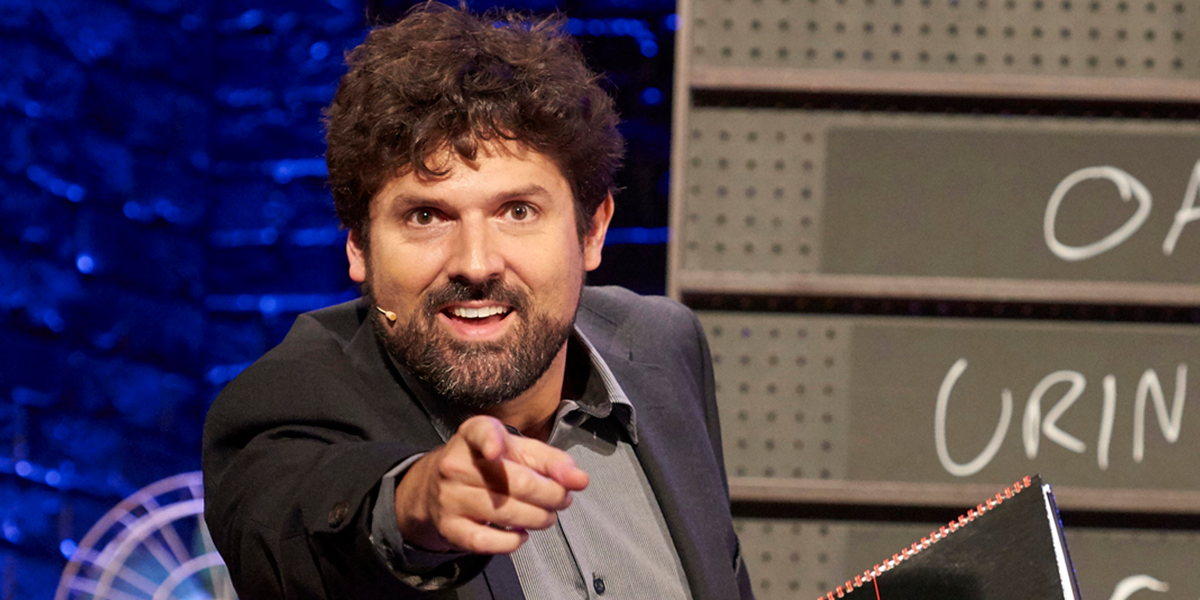 A performer in a bright costume gestures expressively on stage, with vibrant lighting and audience silhouettes in the background.