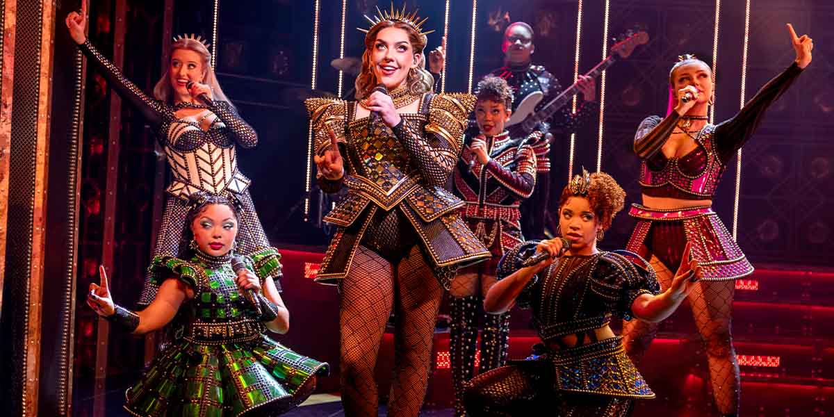 Adrianne Langley as Catherine of Aragon, surrounded by five other performers in vibrant costumes, on stage.