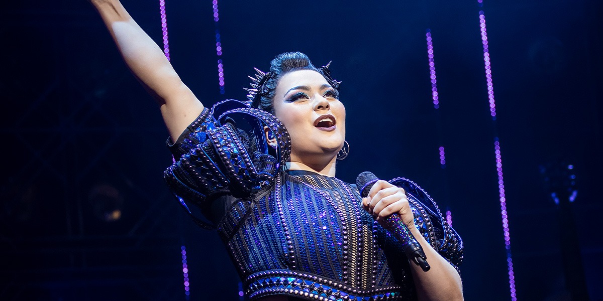 Amelia Kinu Muus as Catherine Parr, wearing a regal costume, stands confidently on stage with dramatic lighting.
