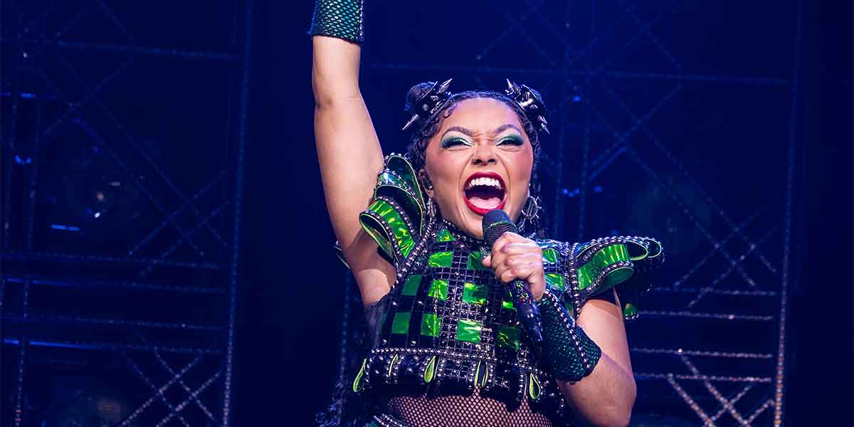 Marisha Morgan as Anne Boleyn, passionately singing with a raised arm, wearing a vibrant green costume.