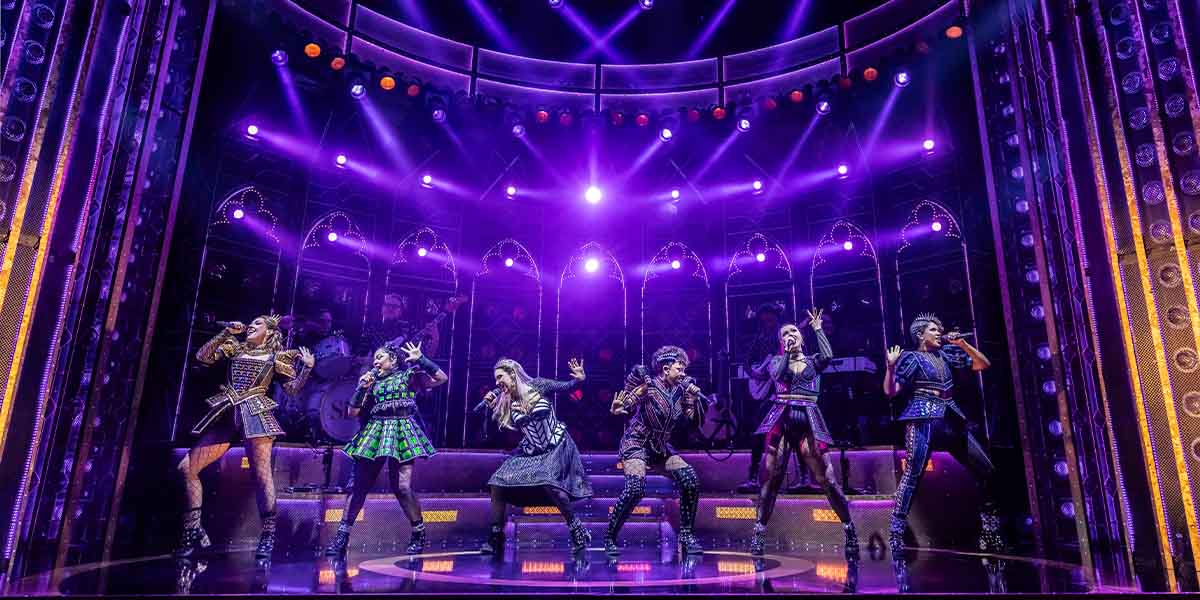 The six cast members of 'Six the Musical' stand together, dressed in vibrant costumes, smiling.