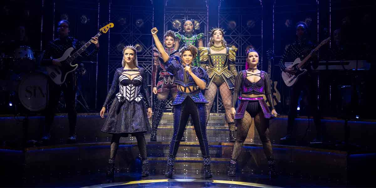 Six performers in vibrant costumes pose confidently on stage, with a band visible in the background.