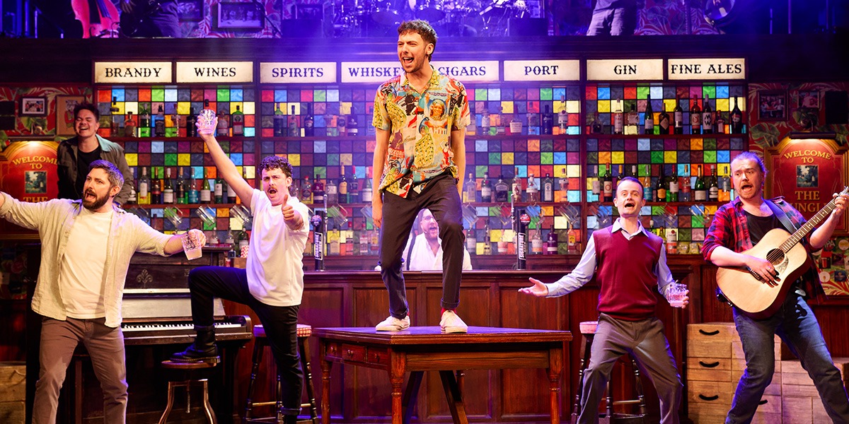 A diverse group of male singers performs energetically on stage, surrounded by musical instruments and warm lighting.