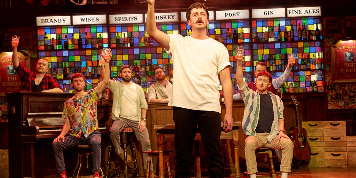 A diverse group of male singers in casual attire perform energetically on stage, surrounded by warm lighting.