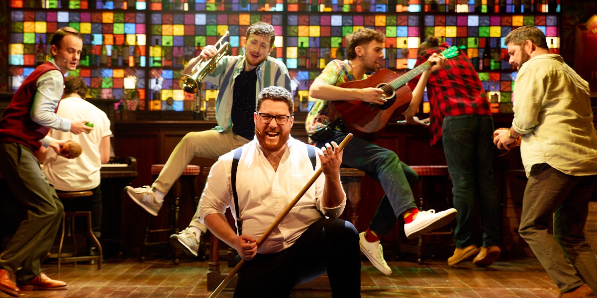 A diverse group of male singers perform energetically on stage, surrounded by vibrant lighting and musical instruments.