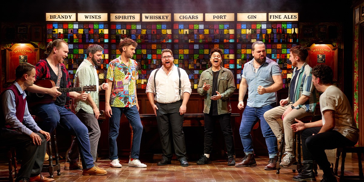 A diverse group of male singers performs energetically on stage, surrounded by vibrant lighting and musical instruments.