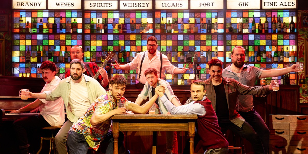 A diverse group of male singers performs energetically on stage, surrounded by warm lighting and wooden decor.