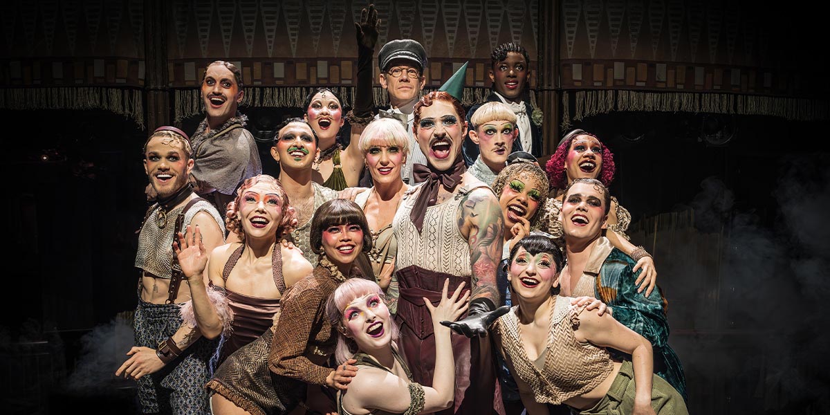 Matt Willis, as Emcee, stands centre stage with performers, vibrant costumes, and dramatic lighting in the background.