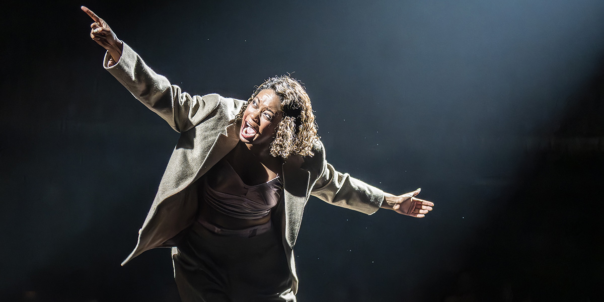 Joy Woods as Sally Bowles passionately performs, wearing a grey coat, with dramatic lighting highlighting her expression.