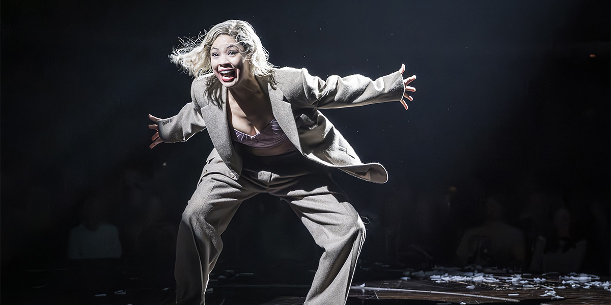 A performer in a sequined costume poses dramatically on stage, surrounded by vibrant stage lights and curtains.