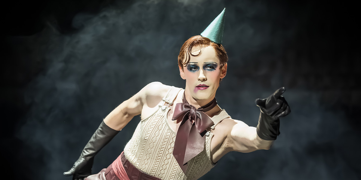 A performer in a sequined costume poses dramatically on stage, with vibrant lighting and a dark background.