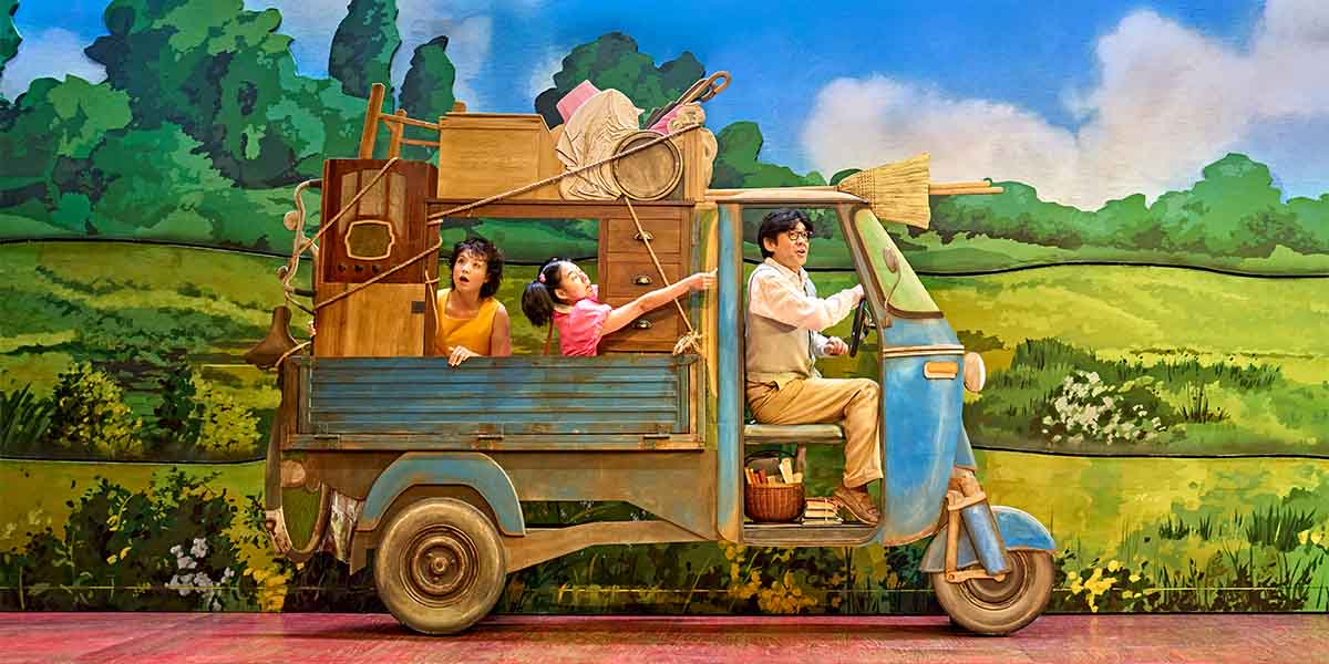 A blue vehicle carries two children and an adult, surrounded by green hills and a bright sky.