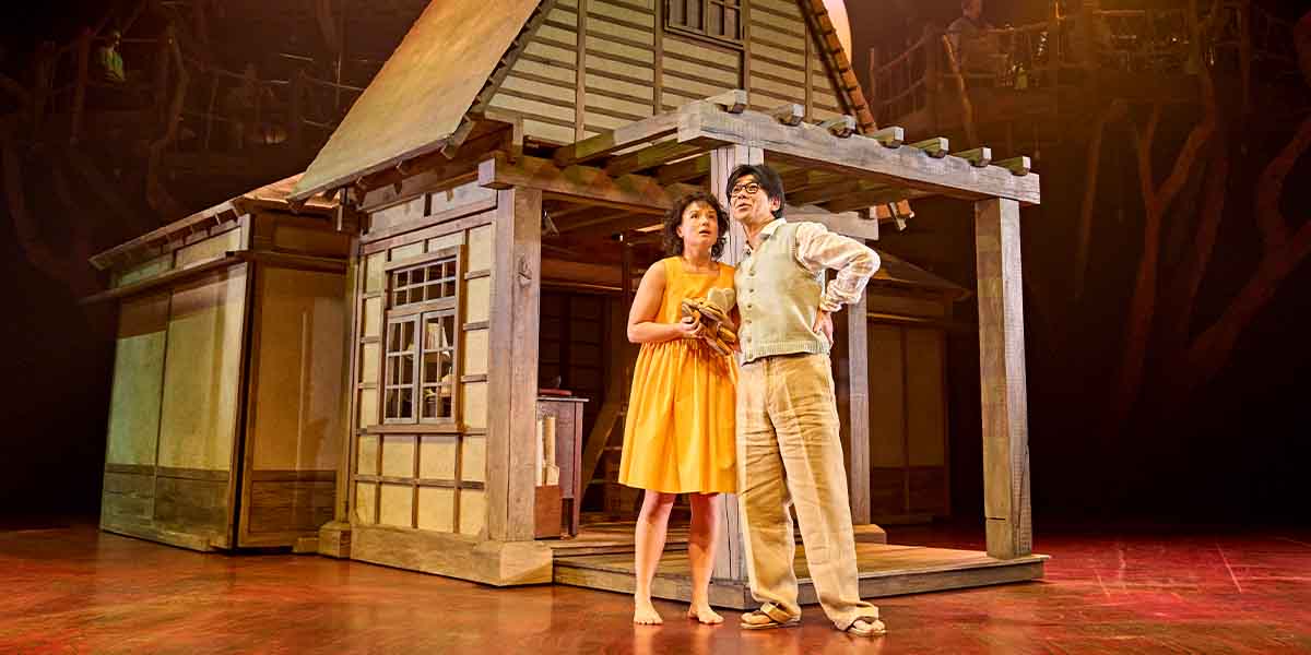 A person in a yellow dress and another in beige stand together by a wooden house backdrop.