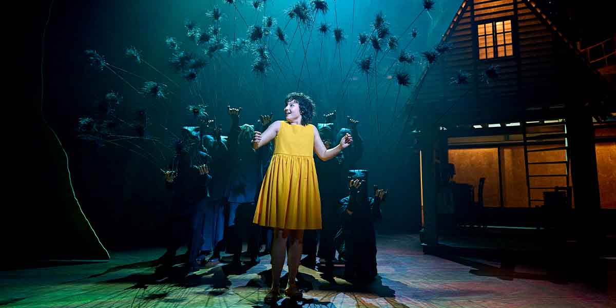 A person in a yellow dress joyfully interacts with shadowy figures holding branches, against a dark stage backdrop.