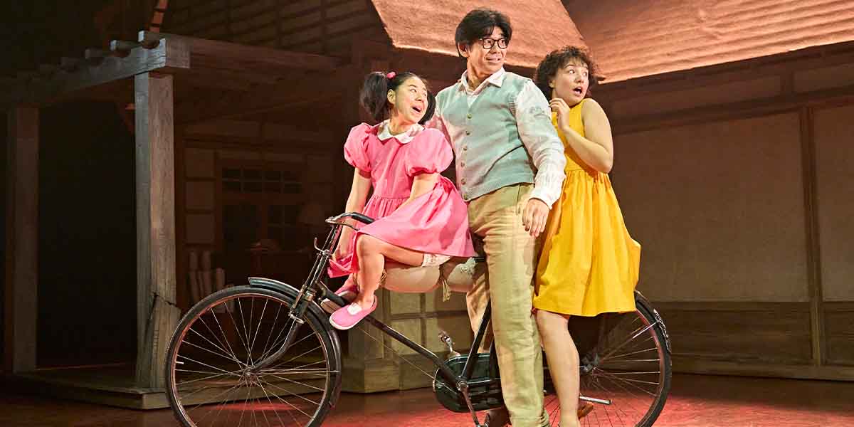 Three characters pose on a bicycle: a person in a pink dress, another in a grey vest, and another in a yellow dress.
