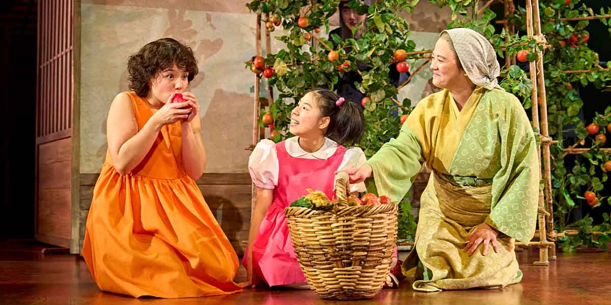 Three characters interact on stage: one in an orange dress holding an apple, another in pink, and another in green.
