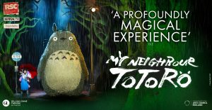 Totoro stands on stage surrounded by green foliage.