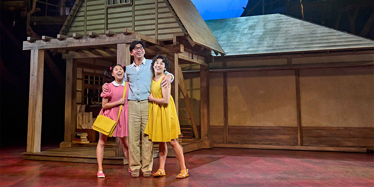 A vibrant scene featuring two children in colourful clothing, joyfully interacting with a large, friendly Totoro under a tree.