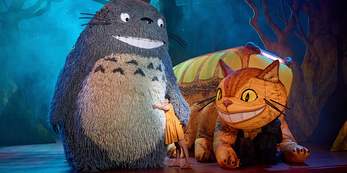 A large, fluffy grey creature with big eyes sits among lush green foliage, surrounded by vibrant flowers.