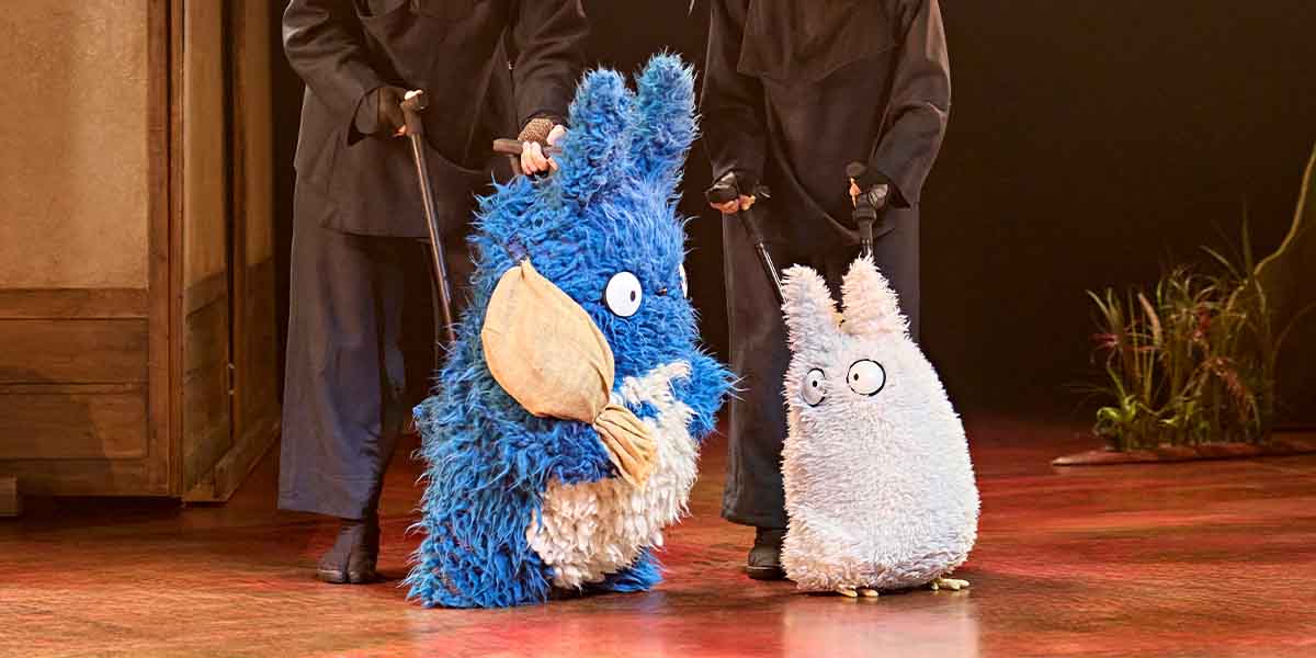 Two large, fluffy puppets, one blue and one white, stand on stage beside two performers in dark costumes.