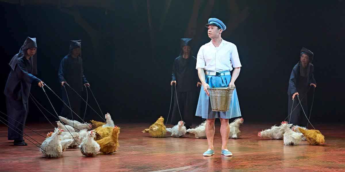 A performer in a white shirt and blue skirt holds a bucket, surrounded by actors with chickens.