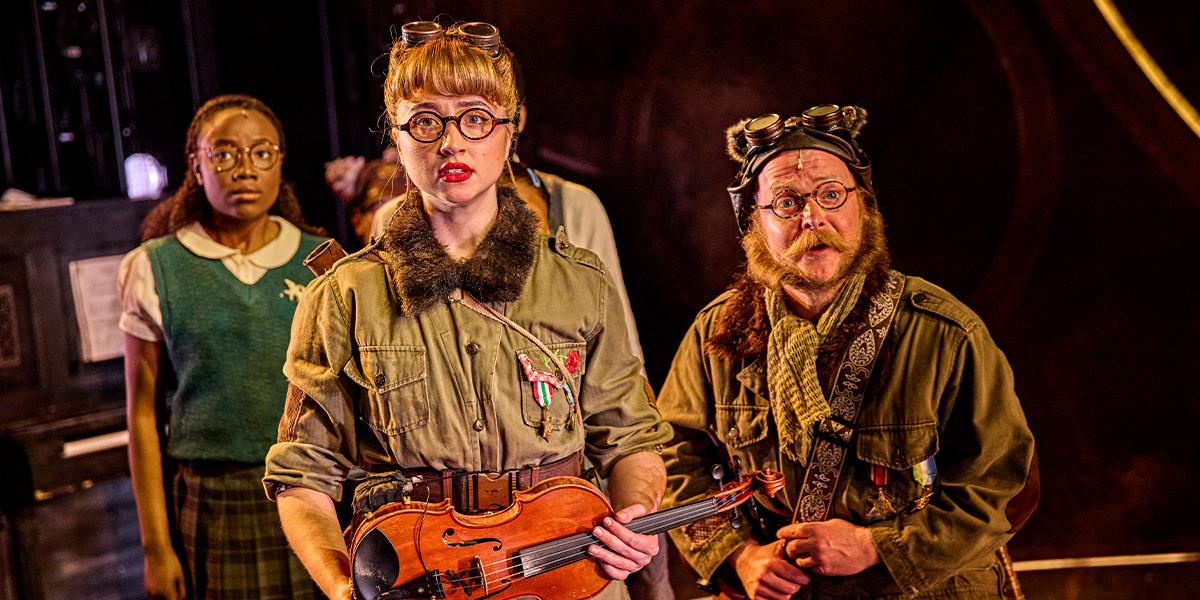 Three people are standing together. Two are wearing googles which they have place on top of their heads and one is playing a ukelele.