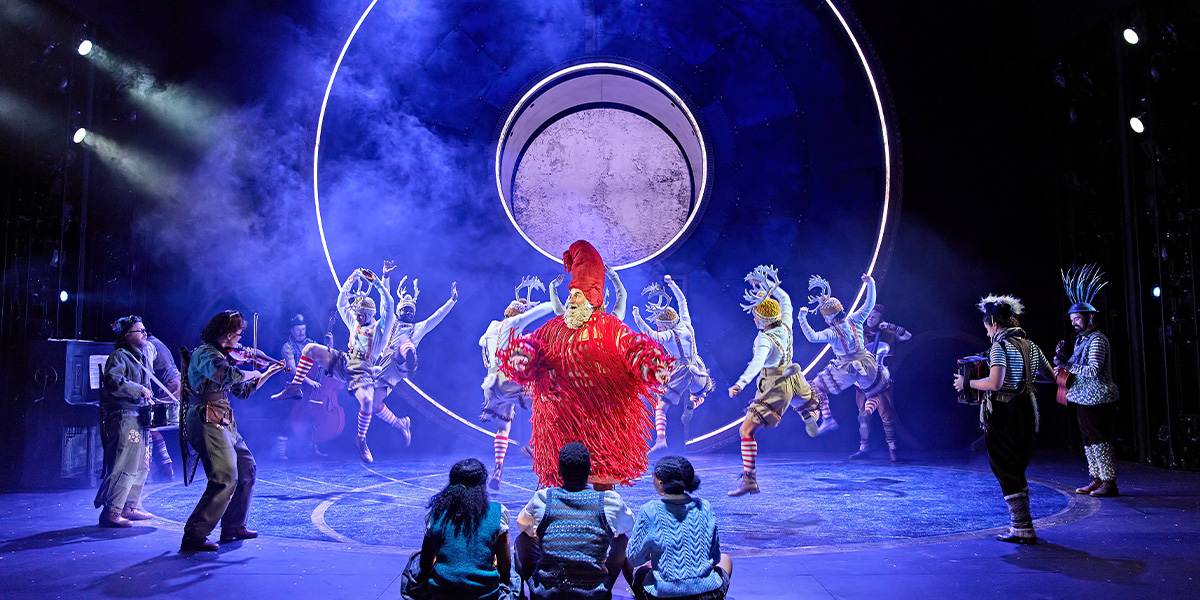 People dance on stage wearing antlers. In the centre is a person wearing a red flowy outfit and pointy red hat.