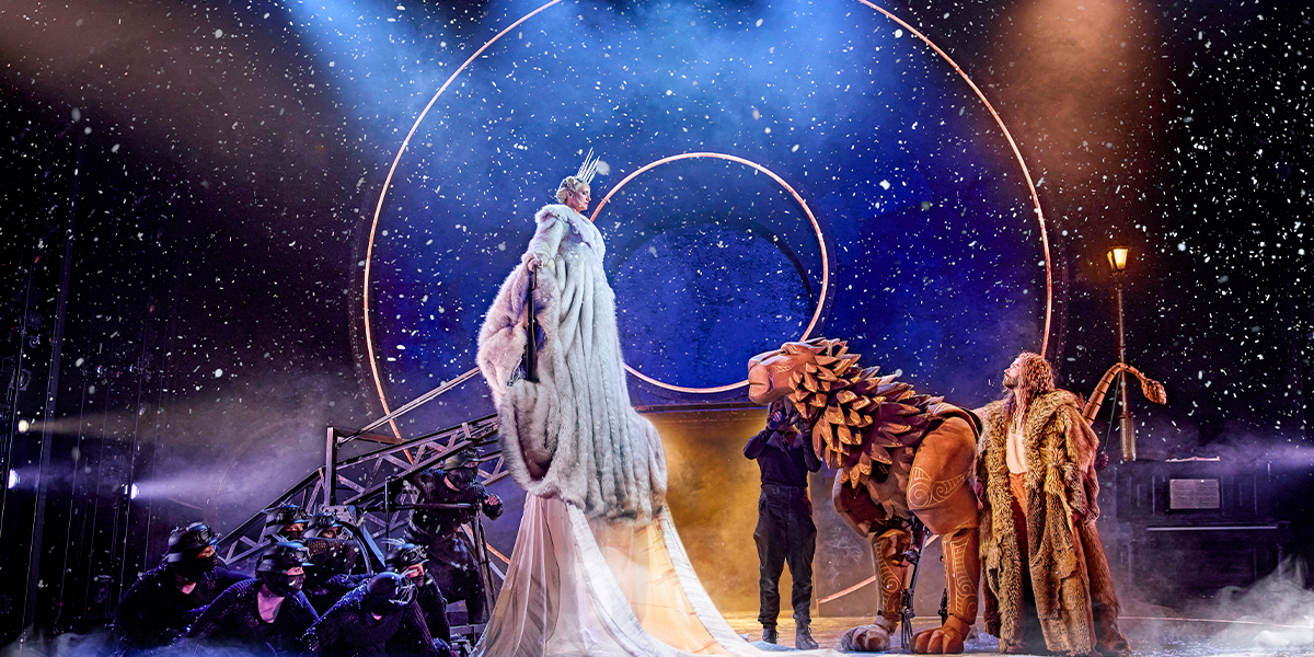 There is a stage with a black backdrop. In the centre, are two light rings; a larger one and smaller one inside it. There is snow in the air. To the right of the image, a woman in long white robes and a white fur coat, and a silver tiara, is lifted off the ground by a wheeled contraption behind her. There is someone wearing a mask behind this contraption and you can only really see there eyes. Her robes are so long, they are covering the snowy ground. To the right of the image is a wooden puppet of a lion. To the left of the lion, closer to the front of the image is a man with long hair wearing a white shirt and auburn trousers with braces. He is wearing a fur coat, similar to the colour of the lion. Both he and the lion are looking up at the woman. There is man behind them, in a black beanie hat.