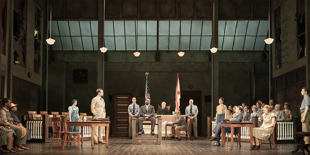 A promotional poster for "To Kill A Mockingbird," featuring the title in bold text against a dark background.