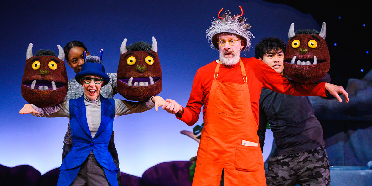 Four performers on stage in a scene. Two are holding large, colourful wild creature puppets, while another is dressed as a character with wild grey hair and an orange jumpsuit, and the fourth performer wears a blue outfit. They are posed playfully against a starry backdrop.
