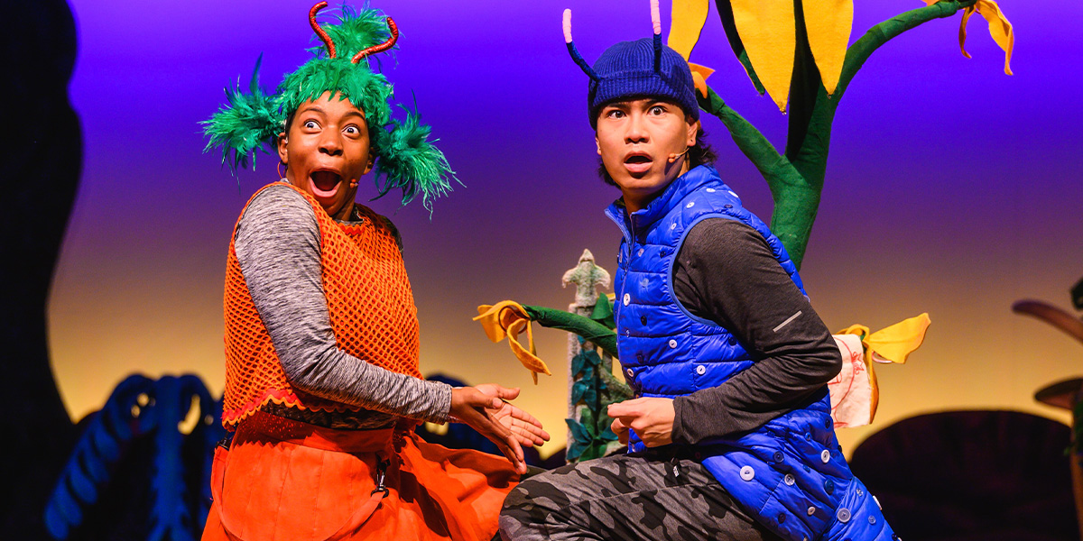 Two actors in colourful costumes performing on stage, one dressed in orange with green hair accessories, the other in a blue vest and hat, both showing expressive facial reactions.