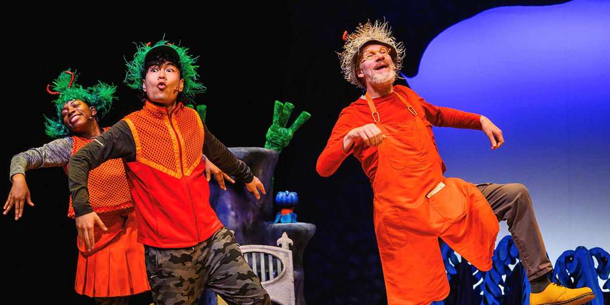 Performers in colorful costumes dancing enthusiastically on stage, with a blue background and a decorative pot.
