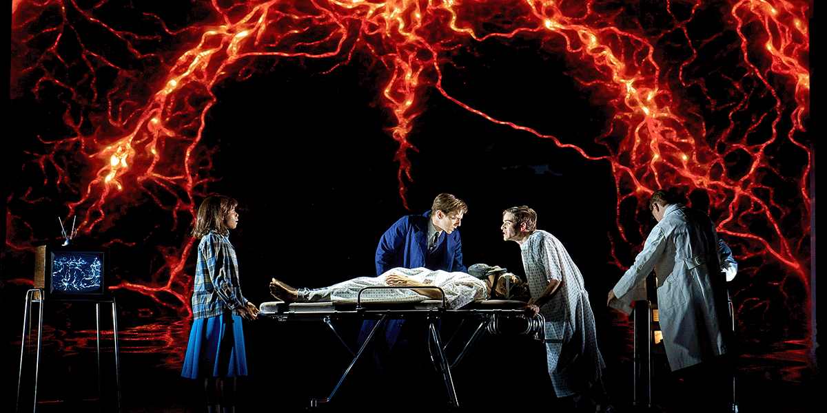 A futuristic and intense stage scene showing characters gathered around a hospital bed with a patient. A glowing red, neural-like pattern fills the backdrop, creating a surreal and ominous atmosphere. Actors are dressed as doctors and a young woman watches nearby.
