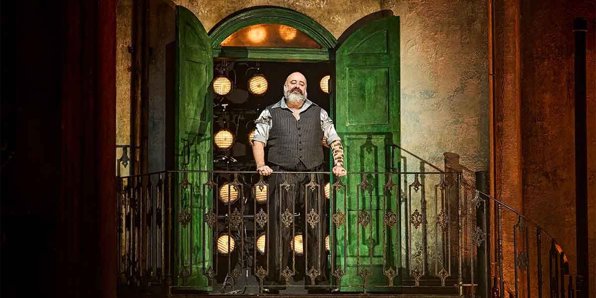 Alastair Parker stands on a balcony with green shutters, smiling, against a backdrop of stage lights.